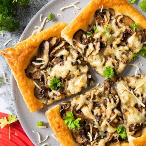 An overhead shot of slices of mushroom tart on a plate, with some shredded cheese and fresh parsley on top; cremini mushrooms are next to the plate.