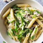 An overhead shot of a serving of pasta with mushrooms and asparagus, with fresh button mushrooms in the background.