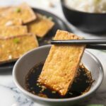 An image showing cripy tofu being dipped in the soy sauce, with white rice on the background.