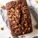 An overhead shot of a chocolate walnut butter cake, ready to be sliced, with walnuts scattered around it.