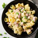 An overhead shot of a serving of ham and pea pasta, garnished with grated cheese and ground black pepper.