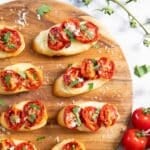 A tray of bruschetta with roasted cherry tomatoes, sprinkled with grated cheese, and some fresh tomatoes next to it.